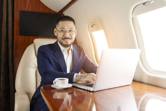 Successful asian businessman in suit and glasses sits in private jet and types on laptop, korean entrepreneur in business clothes flies in airplane, luxury lifestyle.